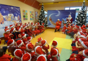 Kilka grup dzieci siedzi na krzesełkach wokół pani dyrektor Marii Królikowskiej, która czyta legendę o świętym Mikołaju w tle dekoracje świąteczne, choinki.